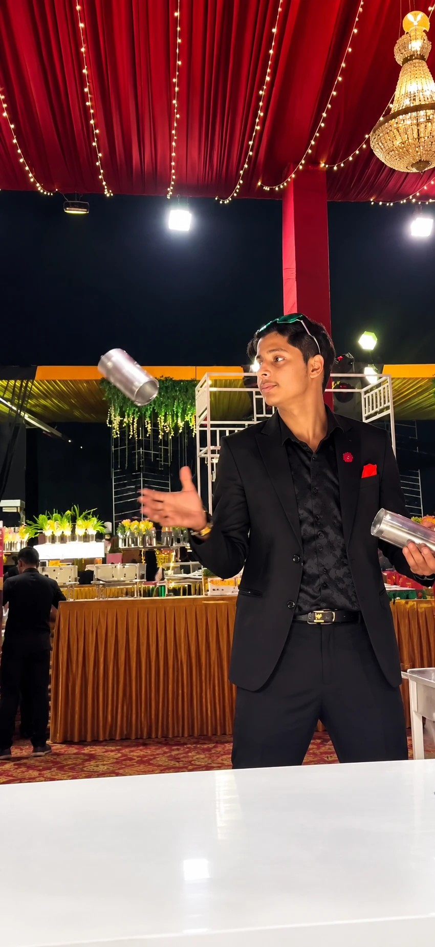 Bartender in a black suit tossing a steel shaker under red drapes at an evening event