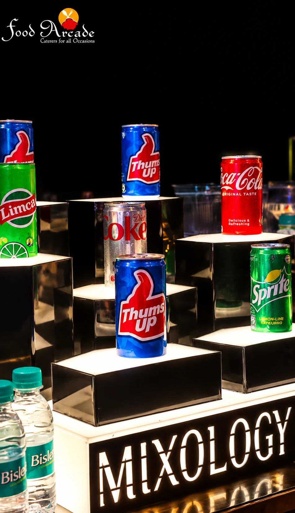 Cans of soft drinks and bottled water arranged on lit steps above a Mixology sign