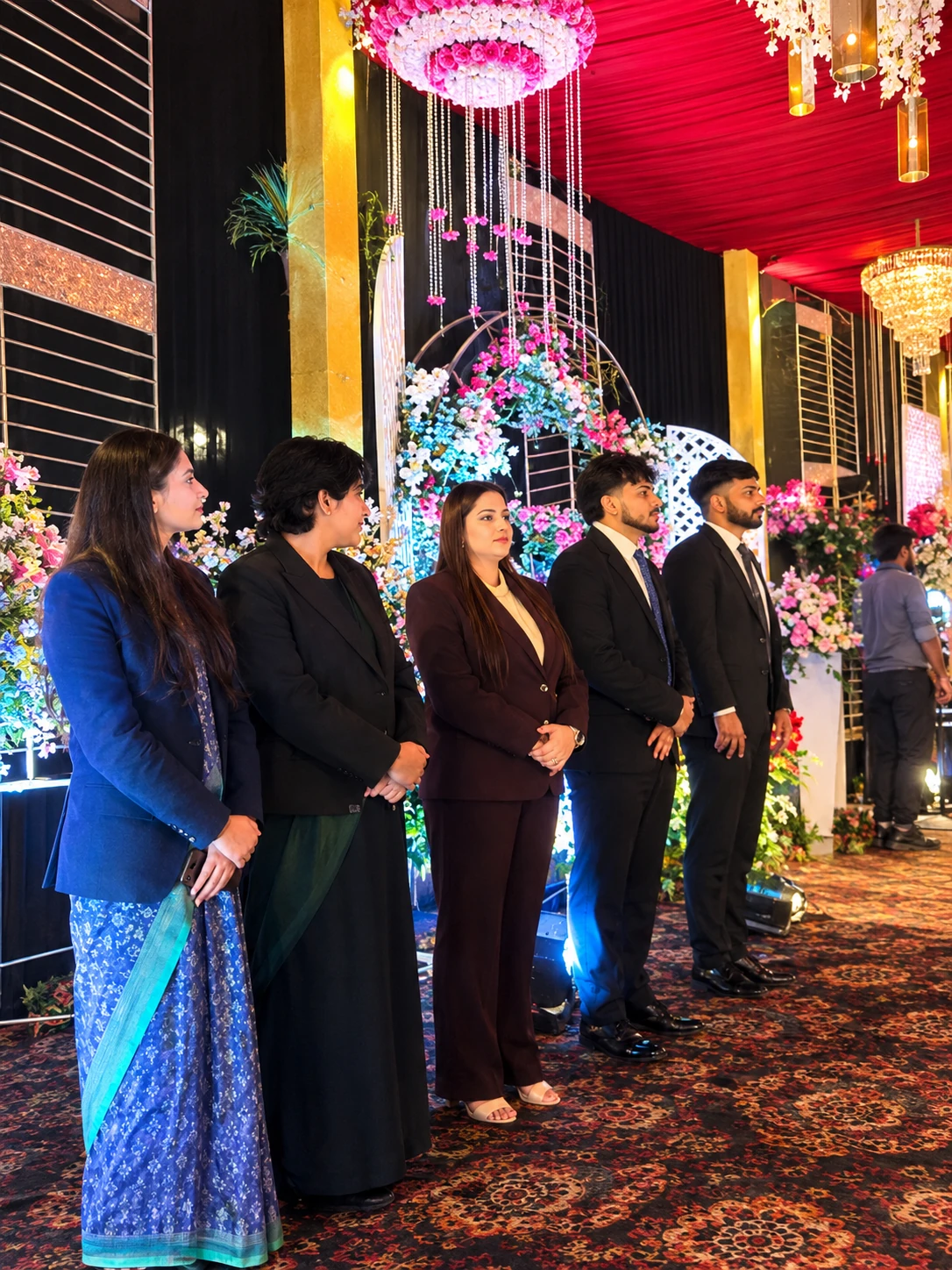 Five team members in formal wear standing in a line in front of a floral stage decoration